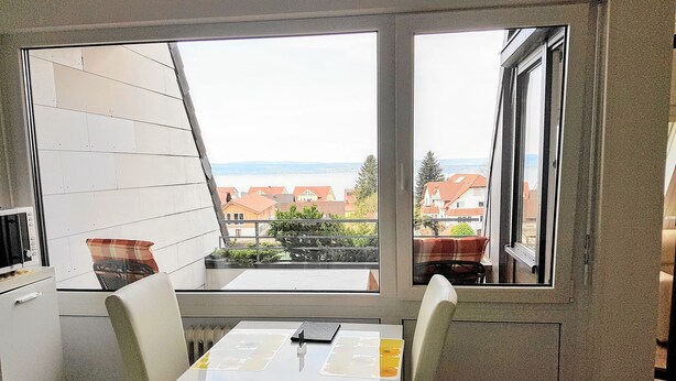 A small kitchen with a table and chairs, a window with a view of houses and trees, and a white cabinet. - Apartment Seeblick (Meersburg)