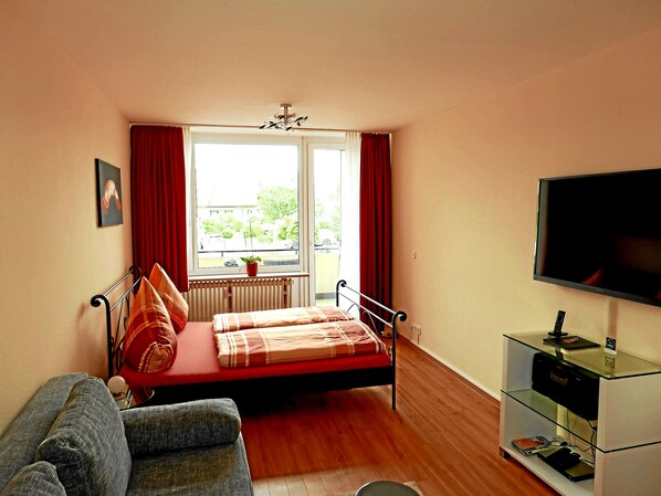 Un dormitorio con cama, televisión, radiador, ventana con cortinas y vistas al exterior.