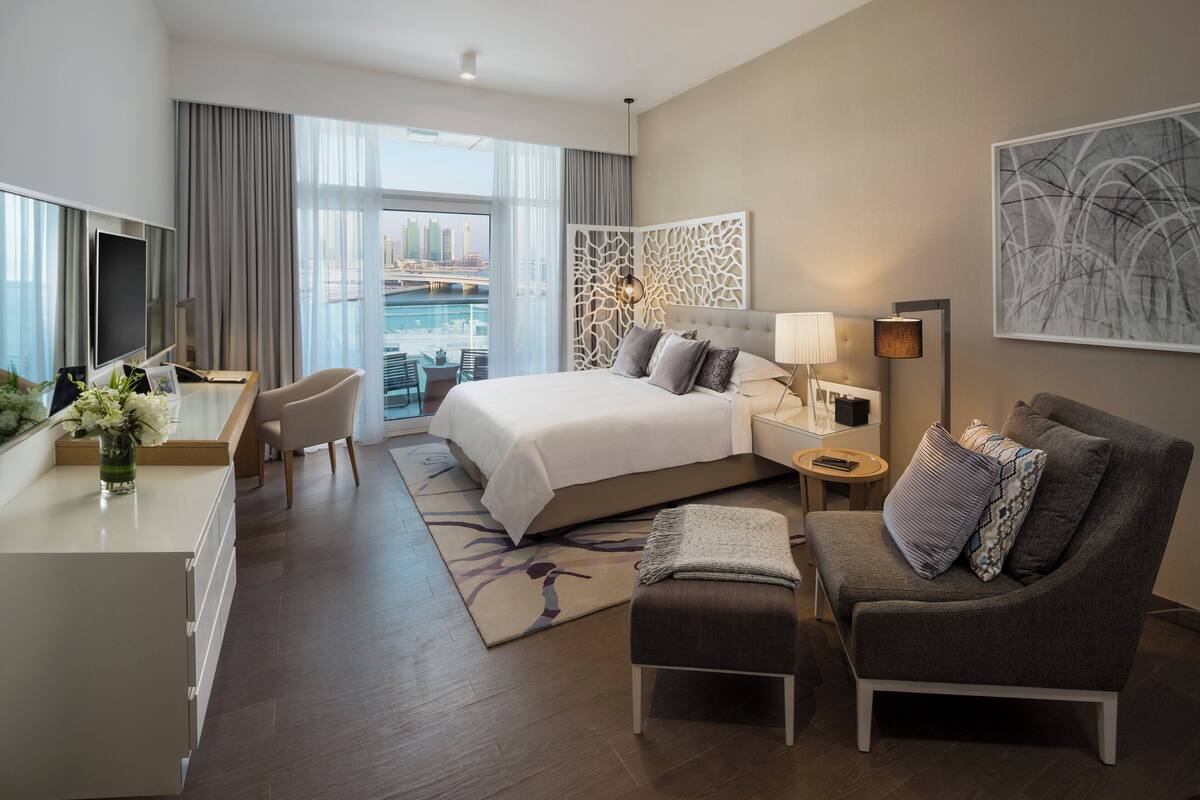 A modern hotel room with a large bed, a desk with a TV, a seating area, and a view of a cityscape.