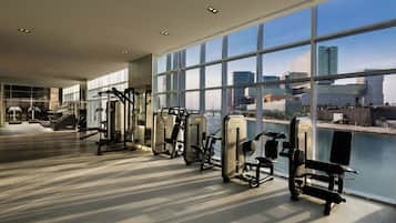 Et fitnesscenter med træningsudstyr og store vinduer med udsigt over vand og bybillede.