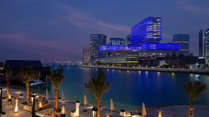 A waterfront outdoor lounge area with seating and palm trees, overlooking a body of water and modern buildings in the background. - Beach Rotana Residences (Abu Dhabi)