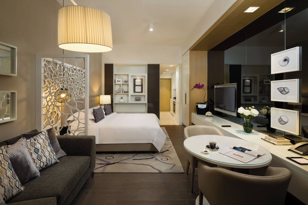 A modern hotel room with a bed, a sofa, a dining table, and a TV.