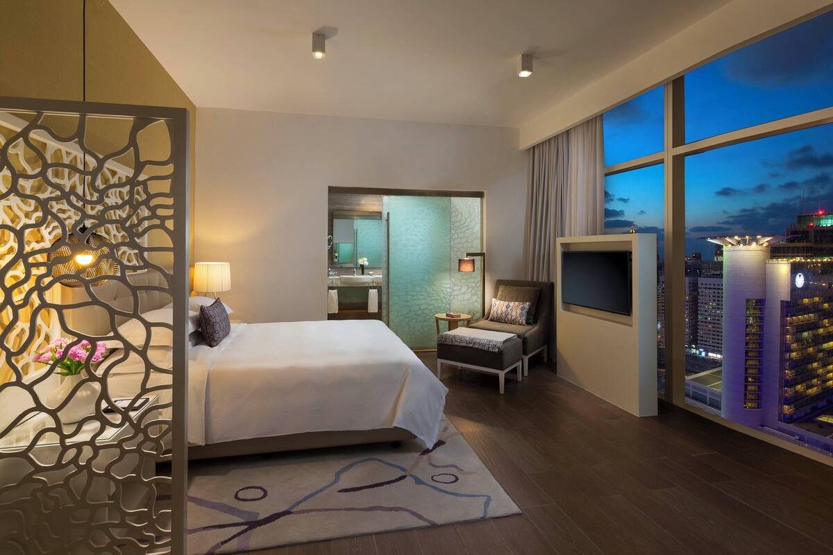 A modern hotel room with a large bed, a seating area, a bathroom, and a city view through large windows.