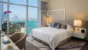 A modern hotel room with a large bed, a desk with a chair, a nightstand, and a view of a cityscape and water. - Beach Rotana Residences (Abu Dhabi)