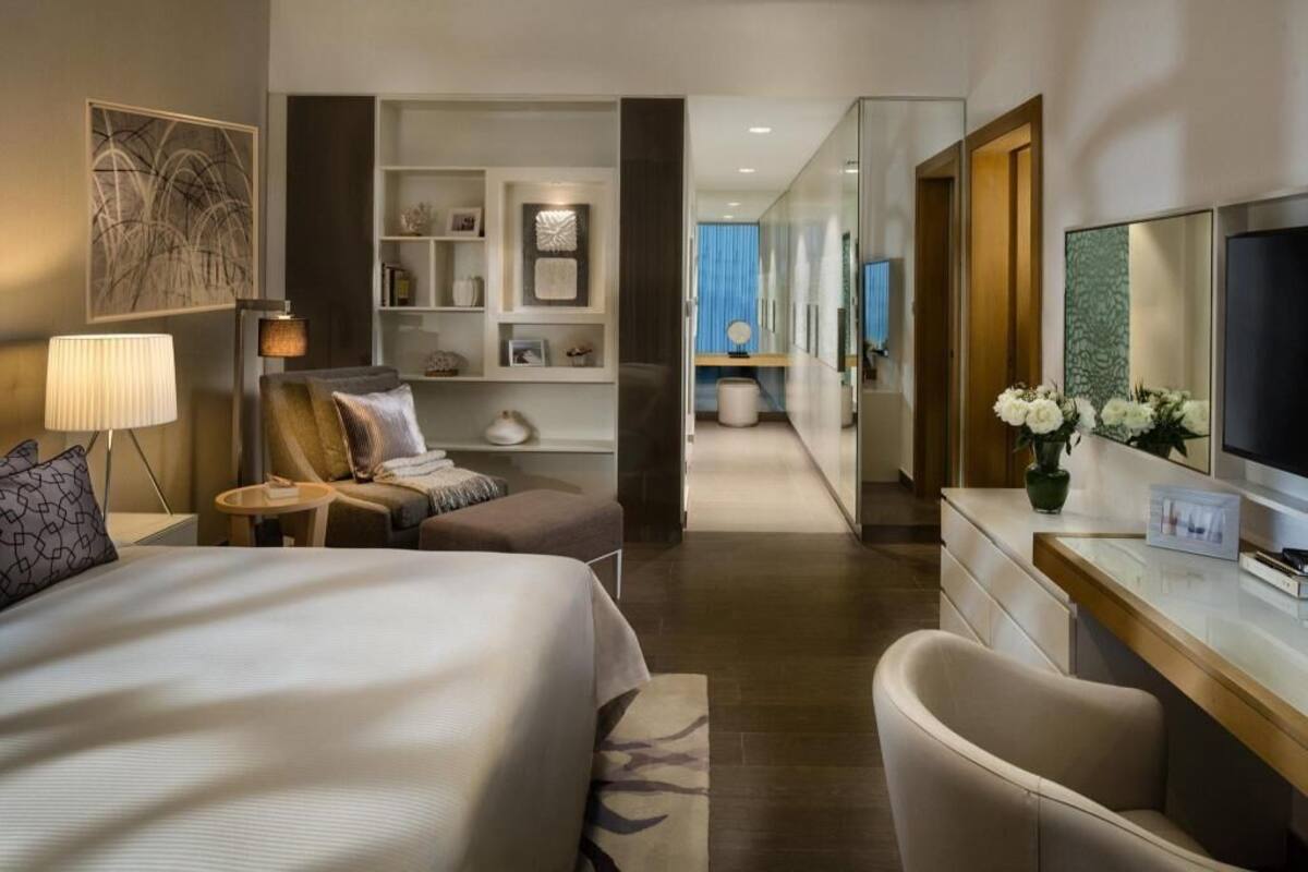 A hotel room with a bed, a desk, a chair, a TV, a lamp, a bookshelf, and a mirror.