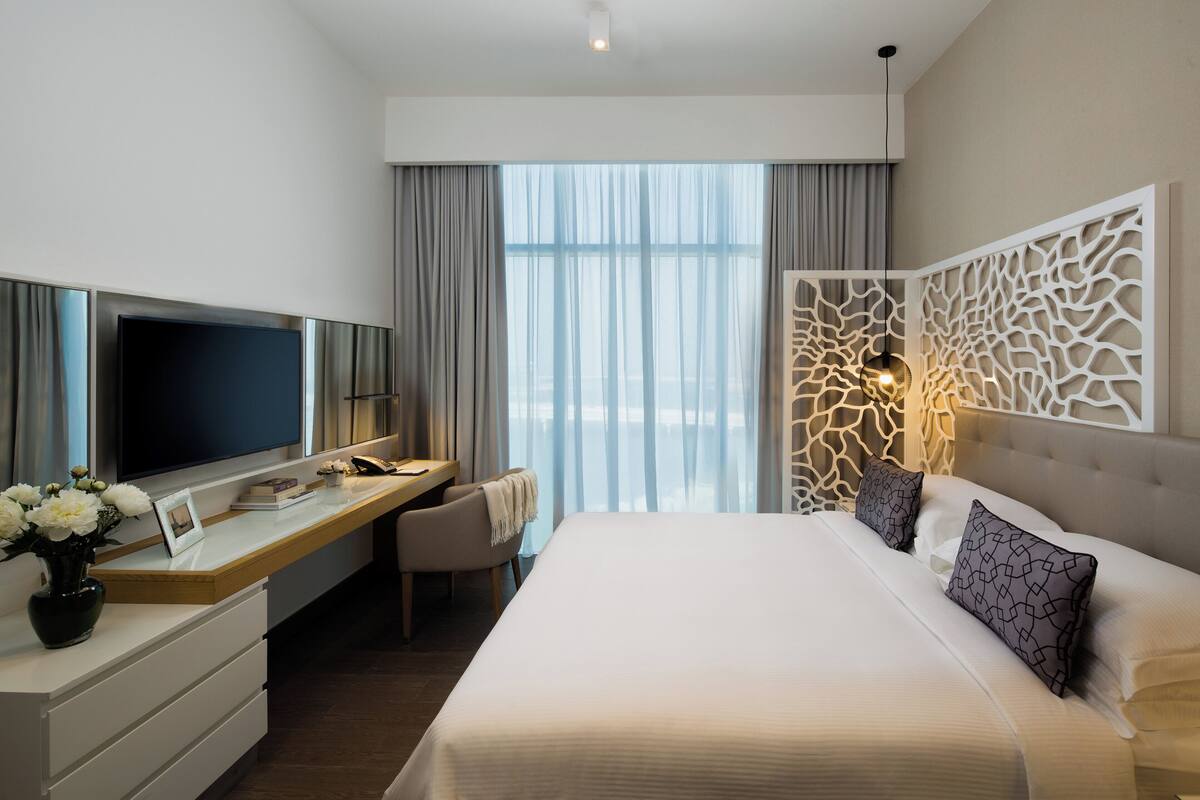 A modern hotel room with a large bed, a desk with a flat-screen TV, a chair, and a decorative wall panel.