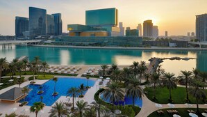 A modern hotel with a large pool, palm trees, and a city skyline in the background. - Beach Rotana Residences (Abu Dhabi)