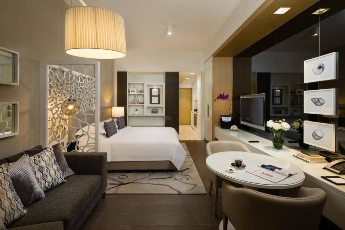 A modern hotel room with a bed, a sofa, a desk with a chair, a TV, and framed pictures on the wall.