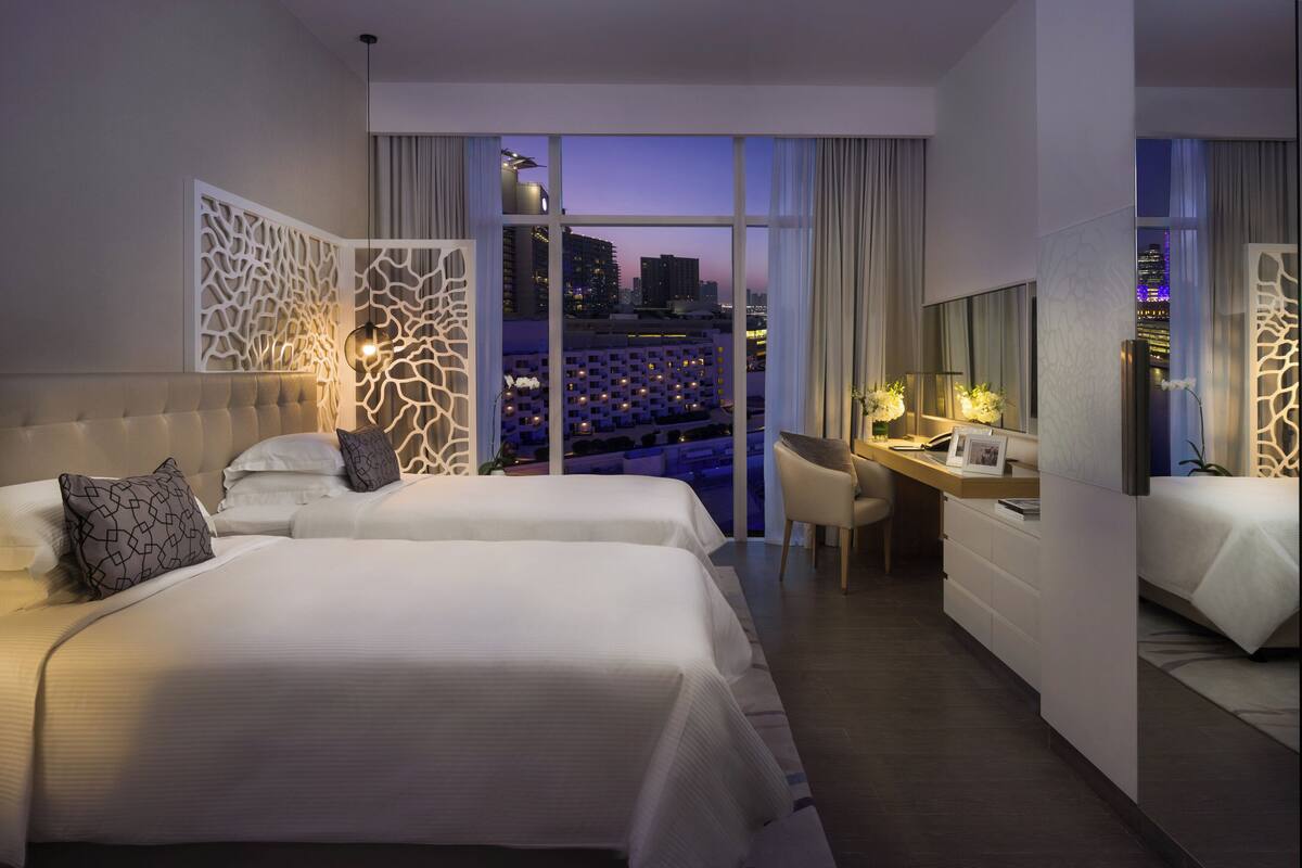 A hotel room with two beds, a desk, and a view of the cityscape at dusk.