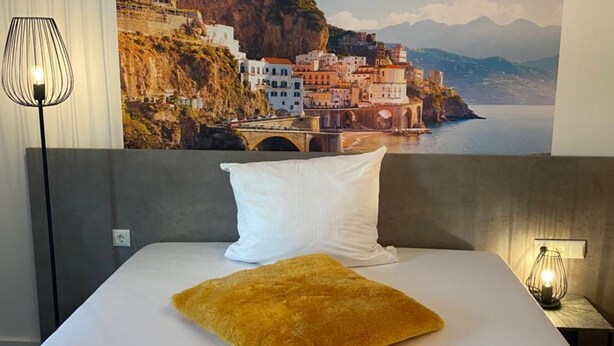 A hotel room with a large bed, a white pillow, a mustard-colored throw, a wall-mounted lamp, and a scenic coastal mural.