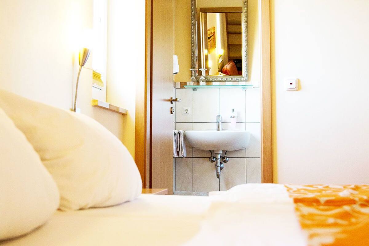 Una habitación de hotel con una cama, almohada blanca, baño con lavabo y espejo, y una toalla colgada en la pared.