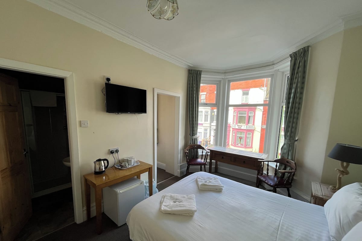 Une chambre d’hôtel avec un lit, un bureau doté d’une bouilloire et de tasses, une télévision, une chaise et une vue sur les bâtiments par la fenêtre.