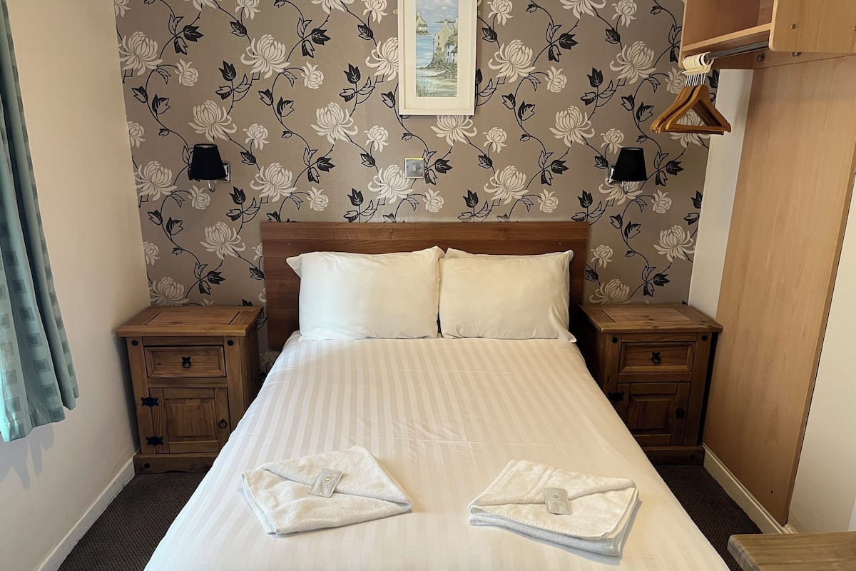 Un lit bien fait, avec du linge de lit blanc, deux oreillers et deux serviettes pliées posées sur le lit. De chaque côté du lit, on trouve des tables de chevet en bois dotées de tiroirs. La pièce est décorée d’un papier peint à motifs floraux et un tableau encadré orne le mur.