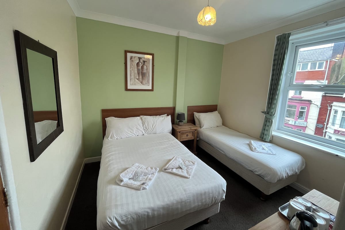 Une chambre d’hôtel avec deux lits, une table de chevet avec une lampe, un tableau encadré au mur et une fenêtre donnant sur des immeubles.
