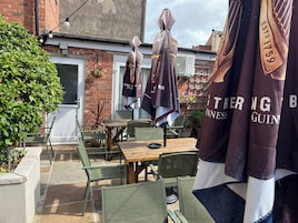 Een terras met tafels, stoelen en parasols, met een bakstenen gebouw op de achtergrond.