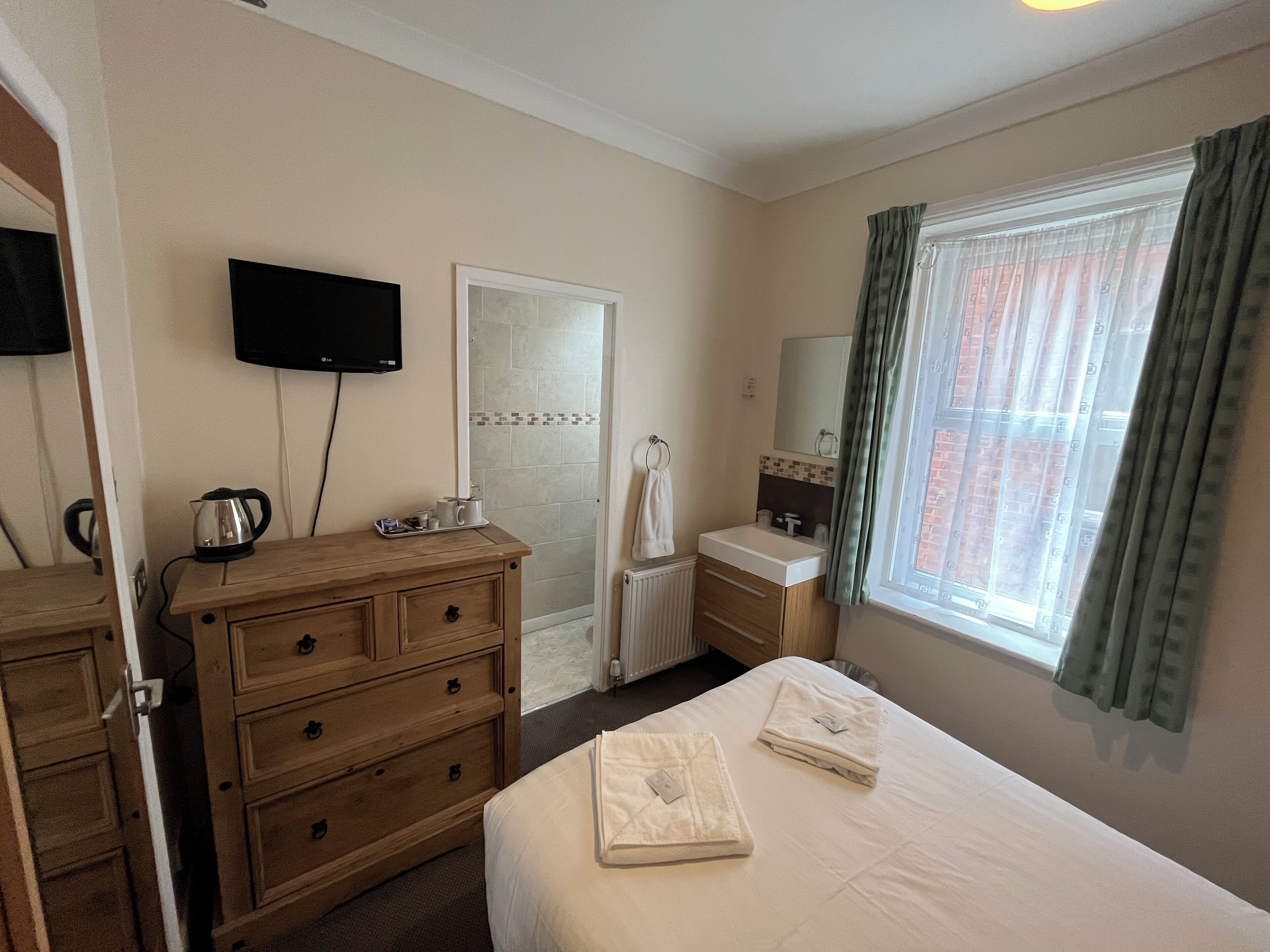 A hotel room with a wooden dresser, a bed with neatly folded towels, a television mounted on the wall, a bathroom with a shower, and a window with curtains.