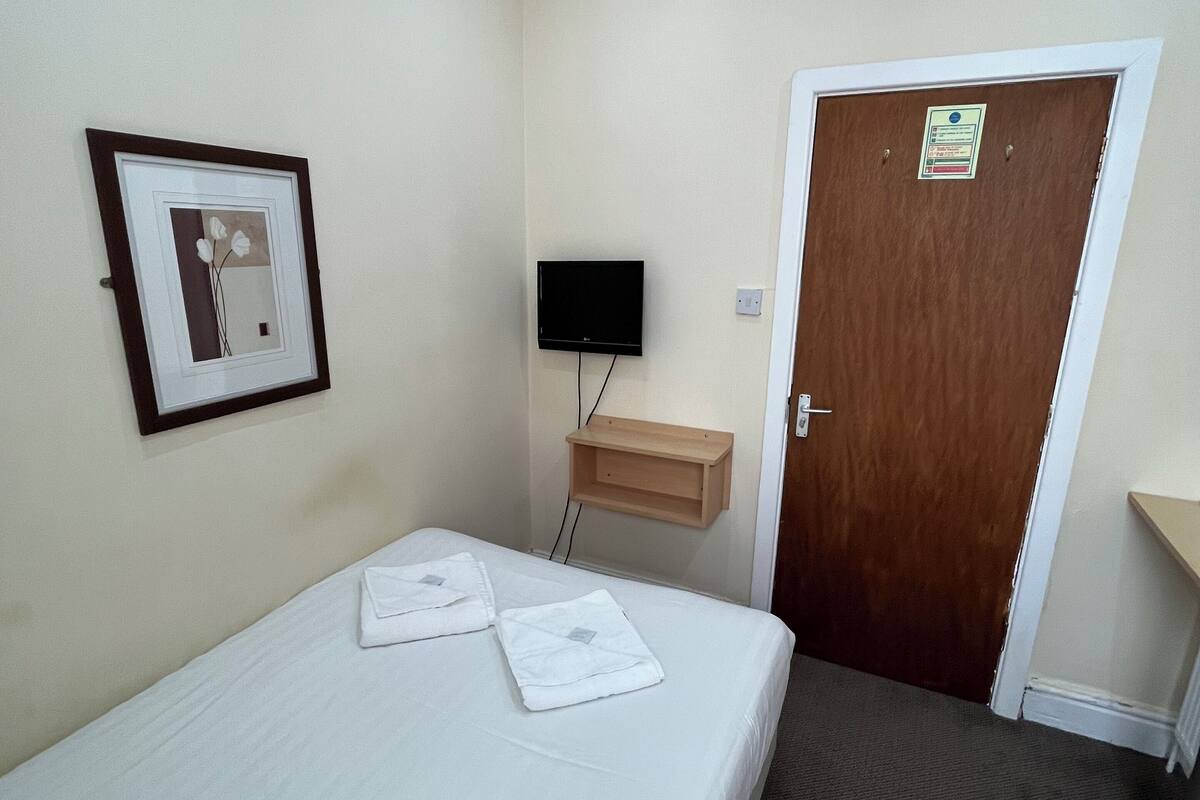 Une chambre d’hôtel avec un lit simple, un téléviseur fixé au mur, une étagère en bois et une porte avec une pancarte.