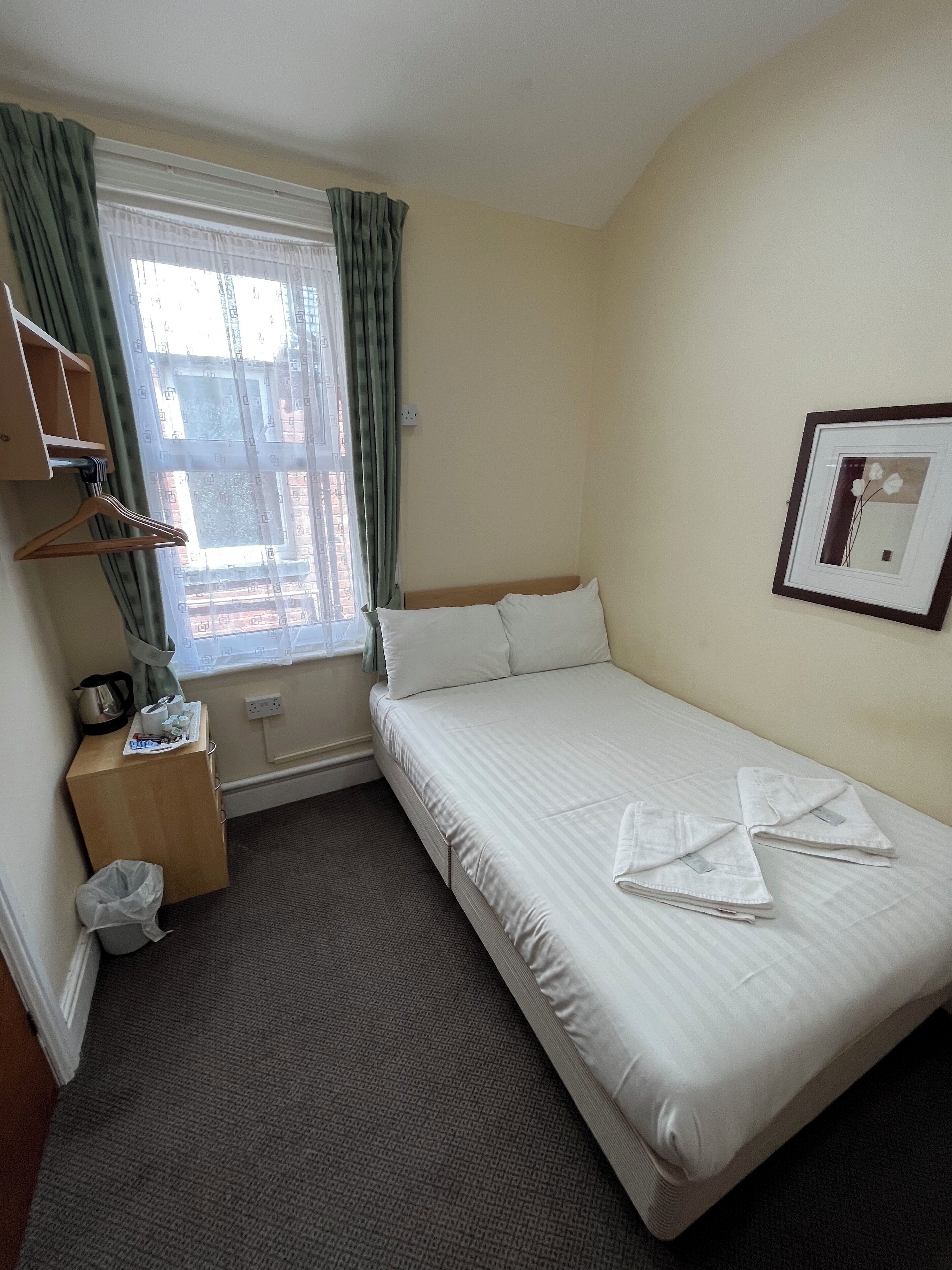 A hotel room with a bed, a small wooden nightstand, a window with curtains, a framed picture on the wall, and a carpeted floor.