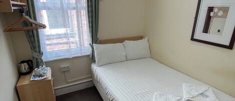 A hotel room with a bed, a small wooden nightstand, a window with curtains, a framed picture on the wall, and a carpeted floor.