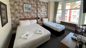 A hotel room with two beds, floral wallpaper, a framed picture, a window with a view of buildings, and a small table with toiletries. - Fairhaven Hotel Central Blackpool (Blackpool)