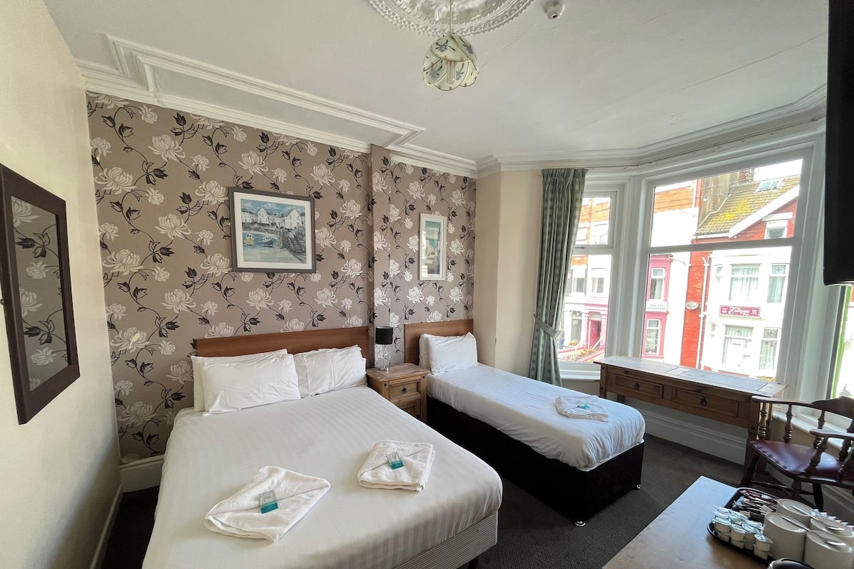 Une chambre d’hôtel avec deux lits, un papier peint à motifs floraux, un tableau encadré, une fenêtre donnant sur des immeubles et une petite table avec des articles de toilette.