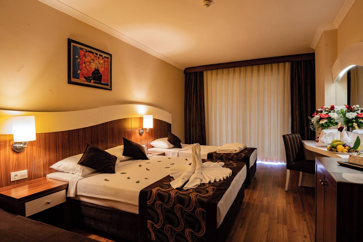 A hotel room with two beds, a desk, and a chair. There is a painting on the wall, a lamp, and a window with curtains.
