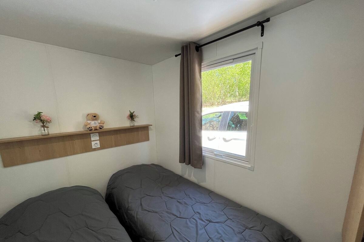 A small room with two beds, a shelf with decorative items, and a window with curtains.