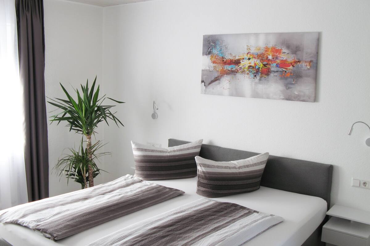 A neatly made bed with striped bedding, a potted plant, and a colorful abstract painting on the wall.