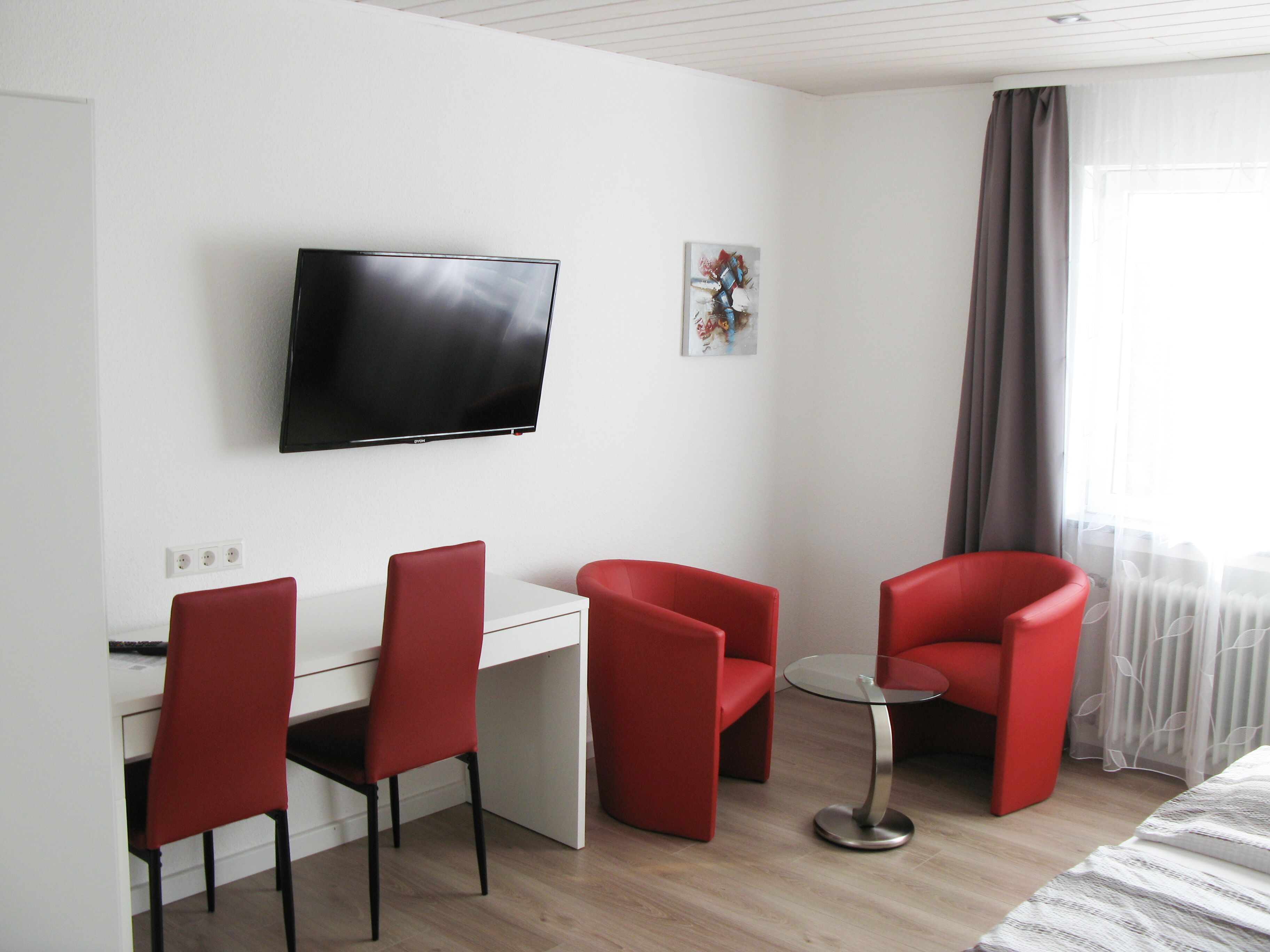 A modern hotel room with a flat-screen TV mounted on the wall, a desk with a chair, and two additional red chairs.