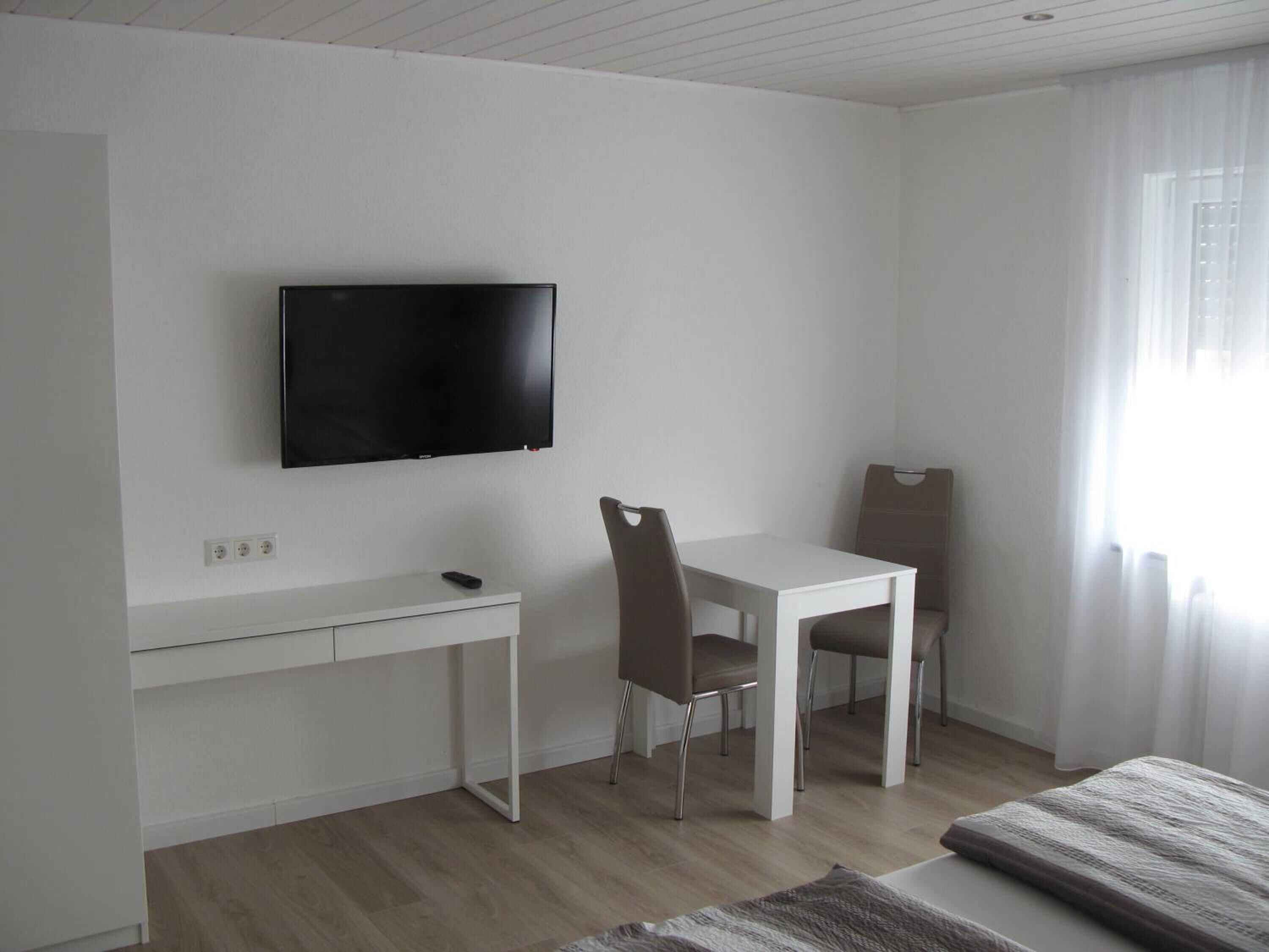 A modern, minimalistic room with a white desk, a chair, a television mounted on the wall, and a large window with curtains.