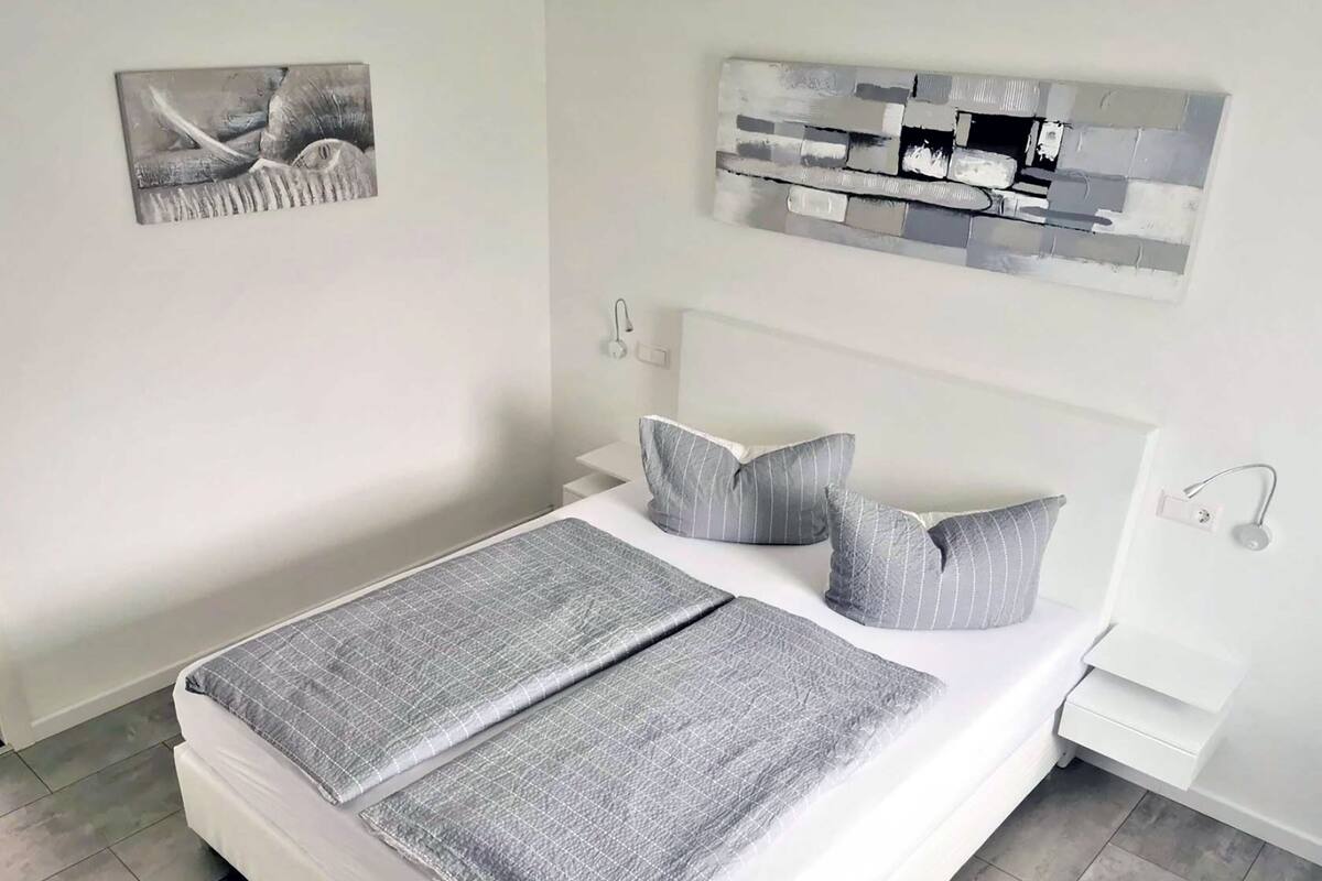 A neatly made bed with a grey checkered blanket and pillows, a white headboard, and a wall-mounted abstract art piece above it.