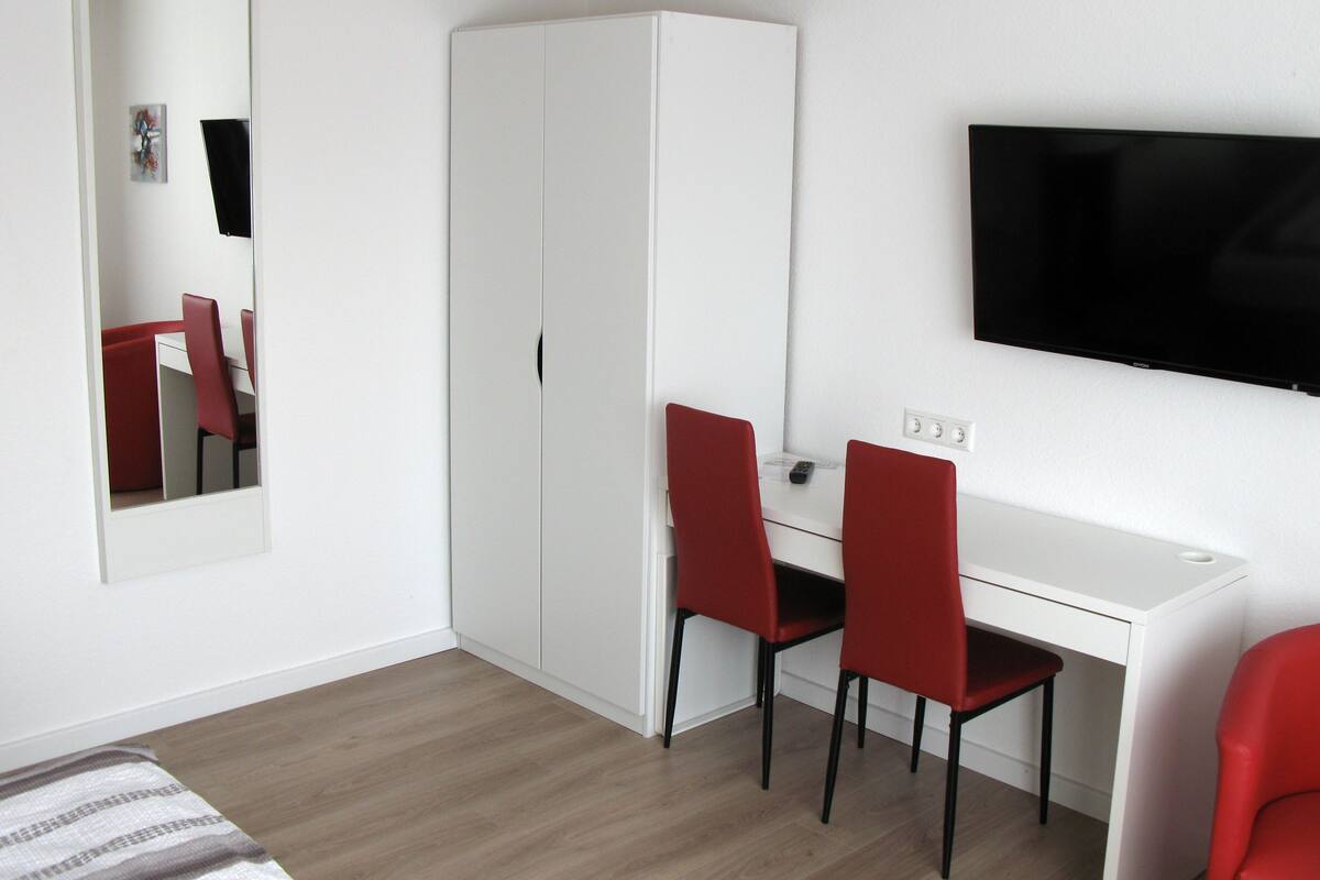 A room with a white desk, two red chairs, a white wardrobe, a flat-screen TV on the wall, and a mirror reflecting the room.