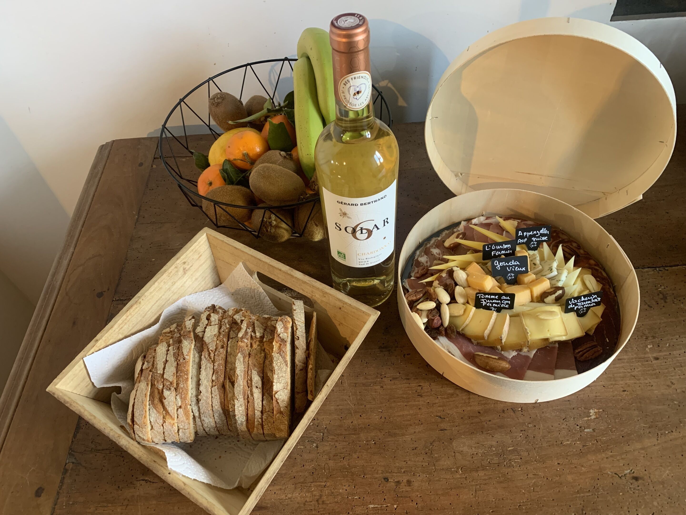 Une table en bois sur laquelle se trouvent une bouteille de vin, un bol de fromage et de noix, un panier de fruits et une miche de pain.