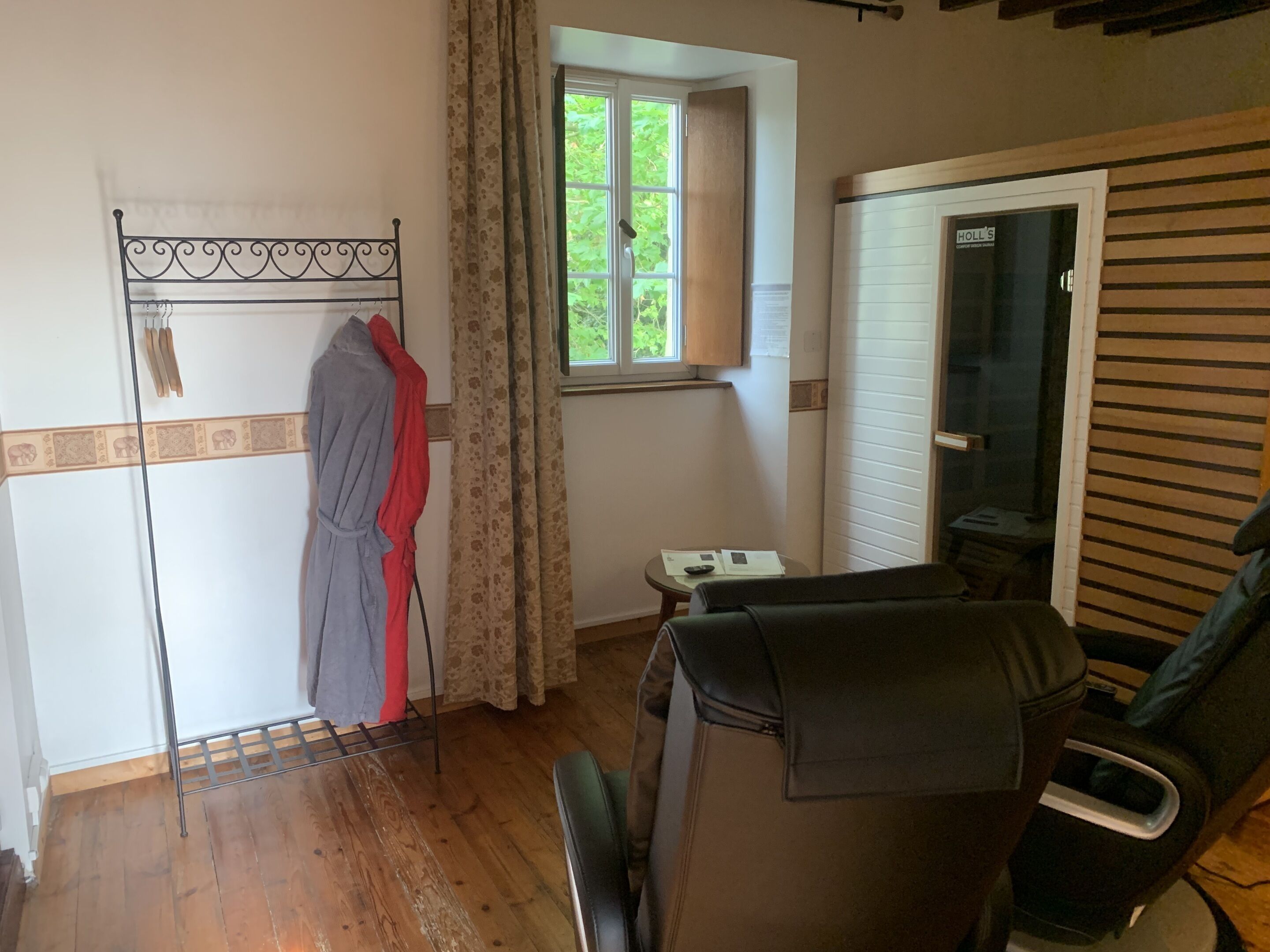 Une pièce avec un plancher en bois, un portant en métal avec des vêtements, une fenêtre avec des rideaux, une petite table avec des papiers et une porte avec une pancarte.