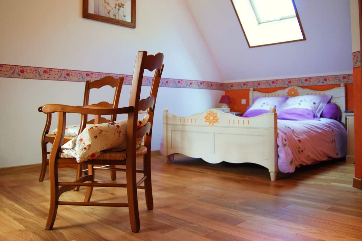Un dormitorio con piso de madera, una cama con ropa de cama floral, una silla de madera, una mesita de noche y un cuadro en la pared.