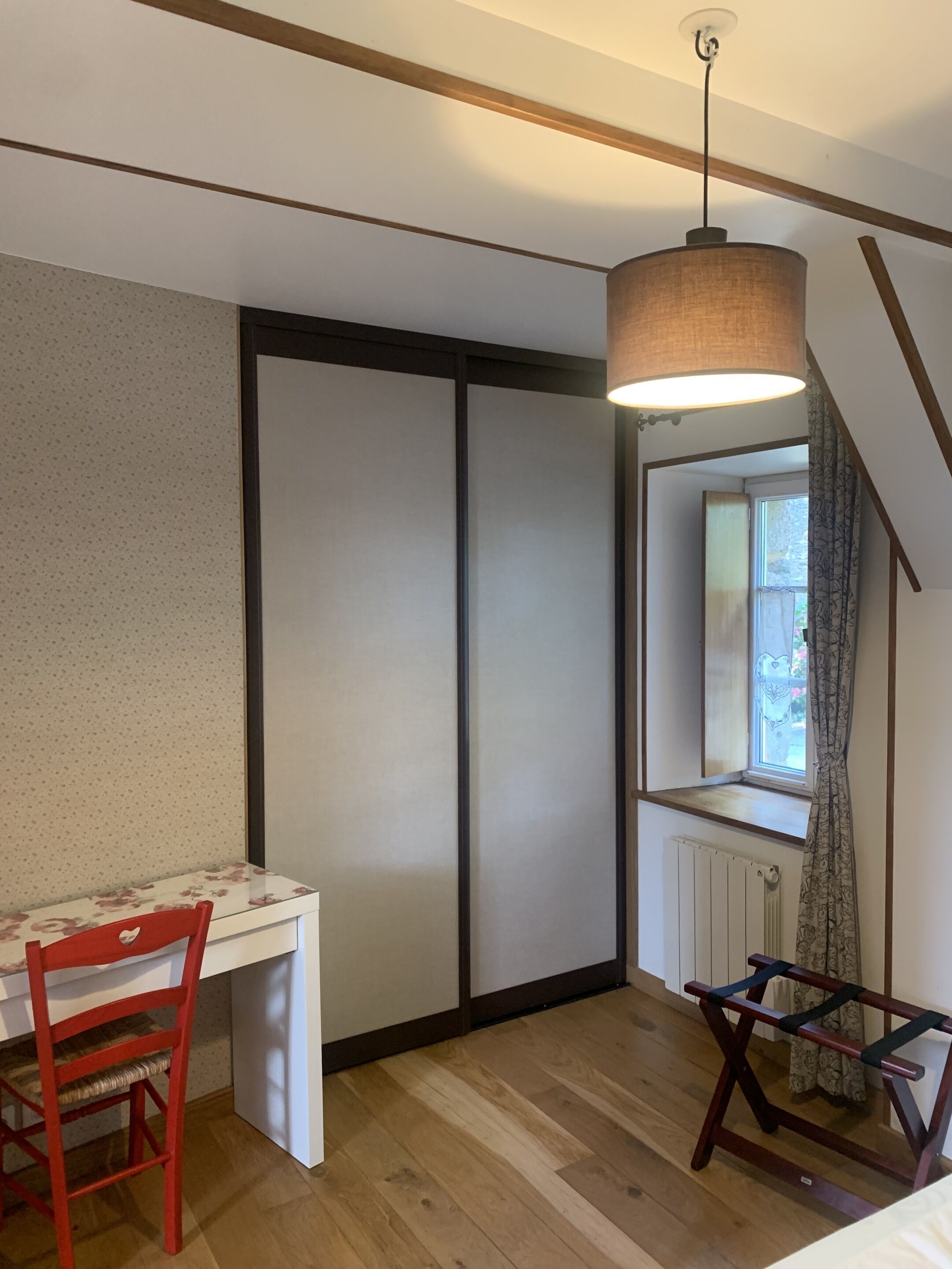 Una habitación con piso de madera, un escritorio blanco con estampado floral, una silla roja, una lámpara colgante y una ventana con vista a follaje exterior.