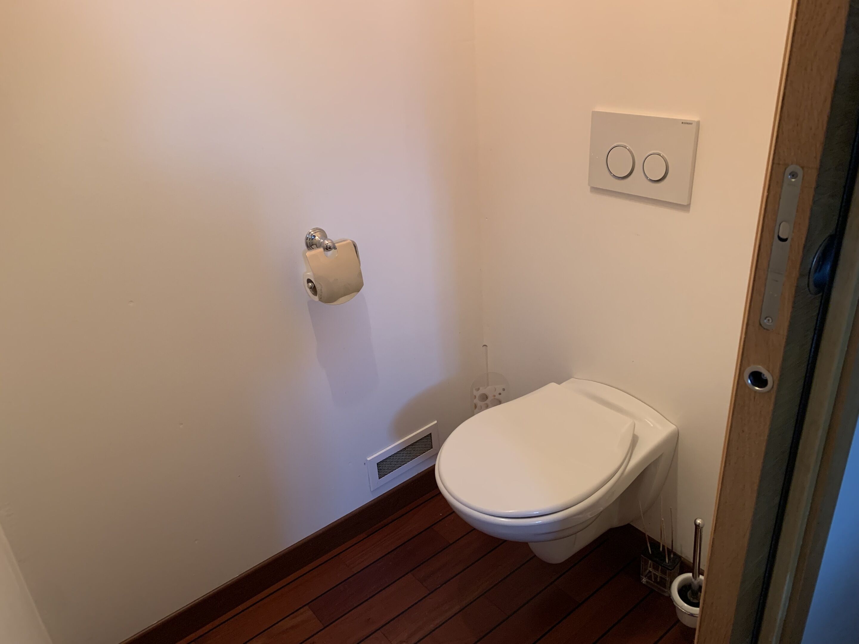 Ein kleines, sauberes Badezimmer mit Toilette, Papierhalter, Lüftung und Lichtschalter an der Wand.
