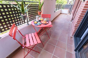 Un balcón con mobiliario rojo, una planta en maceta y una mesa con un croissant y jugo de naranja.