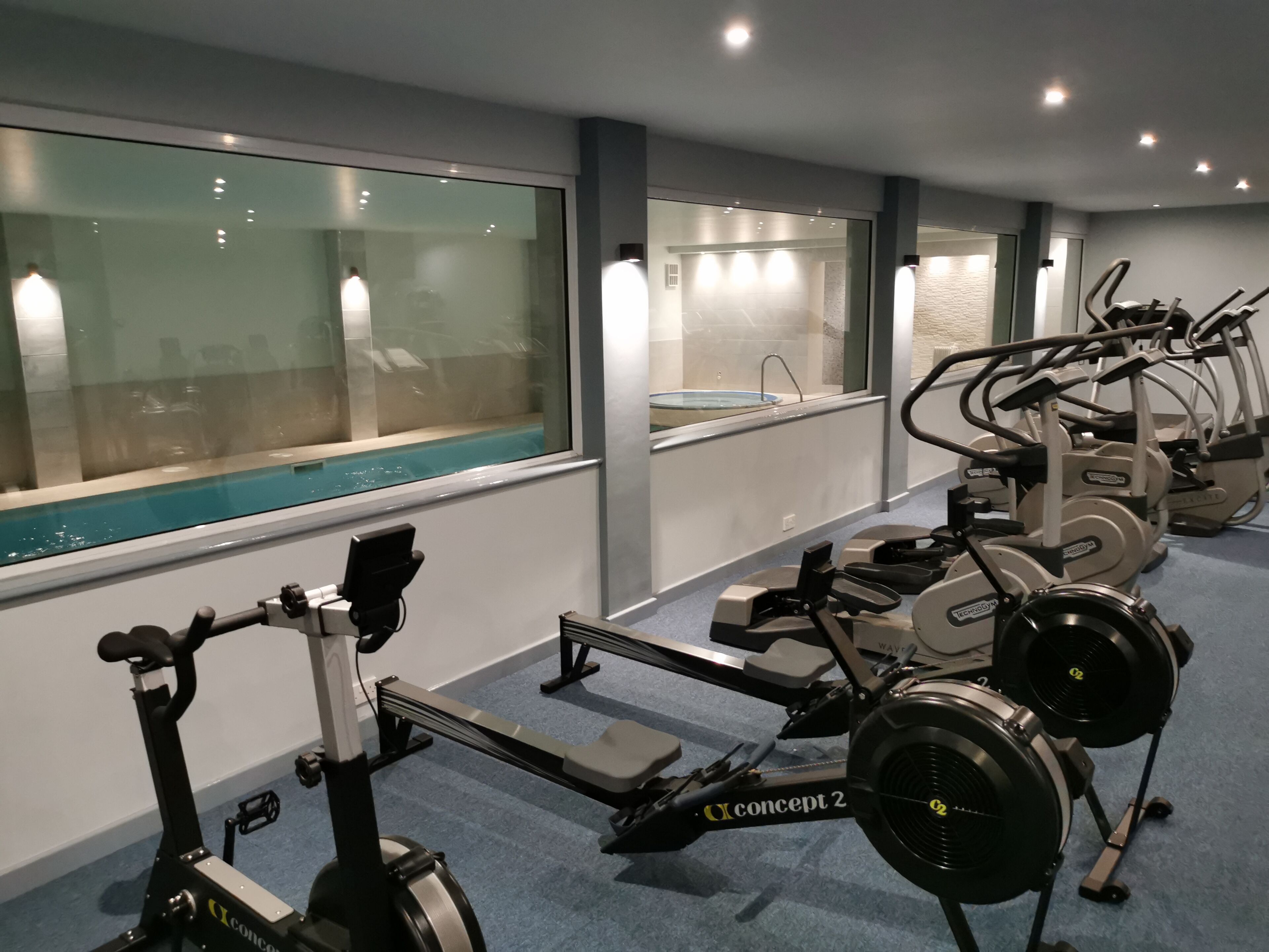Ein Fitnessraum mit Cardiogeräten, einem Rudergerät und Blick auf einen Pool durch große Fenster.