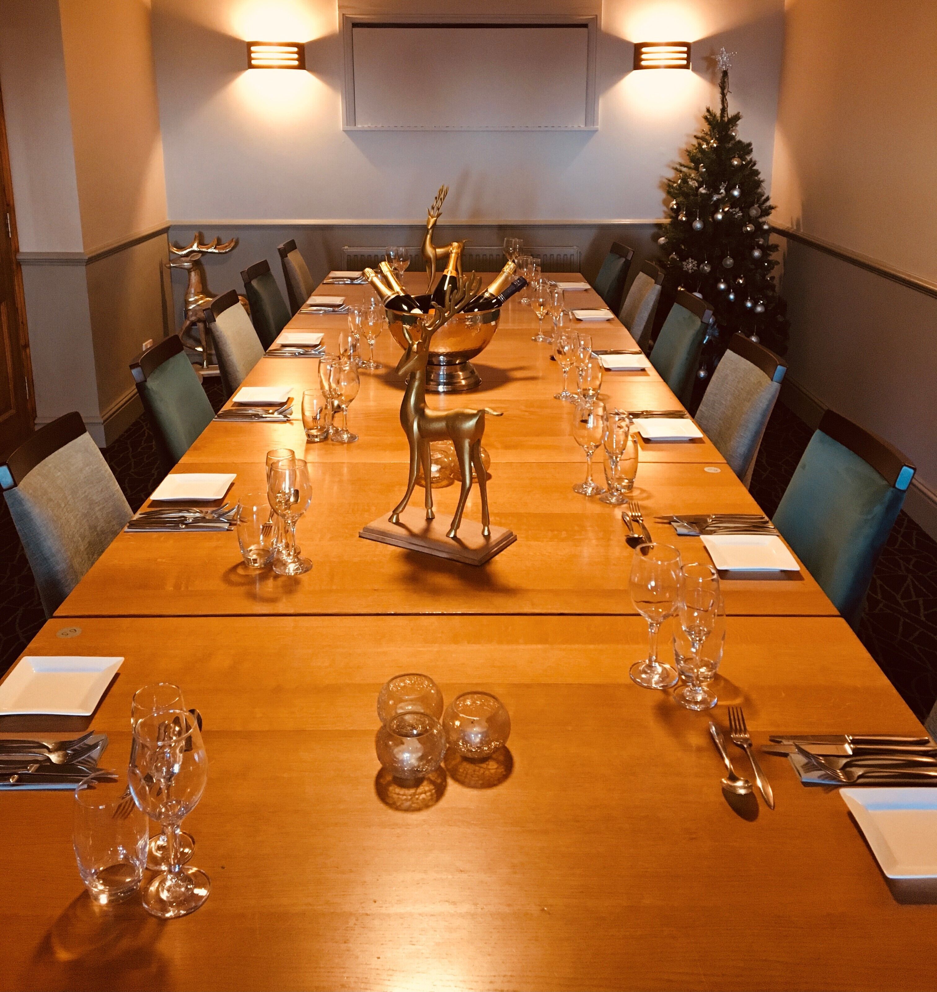 Una larga mesa de comedor preparada para una comida formal, con varias copas de vino, un centro de mesa con una escultura dorada de reno y un árbol de Navidad al fondo.