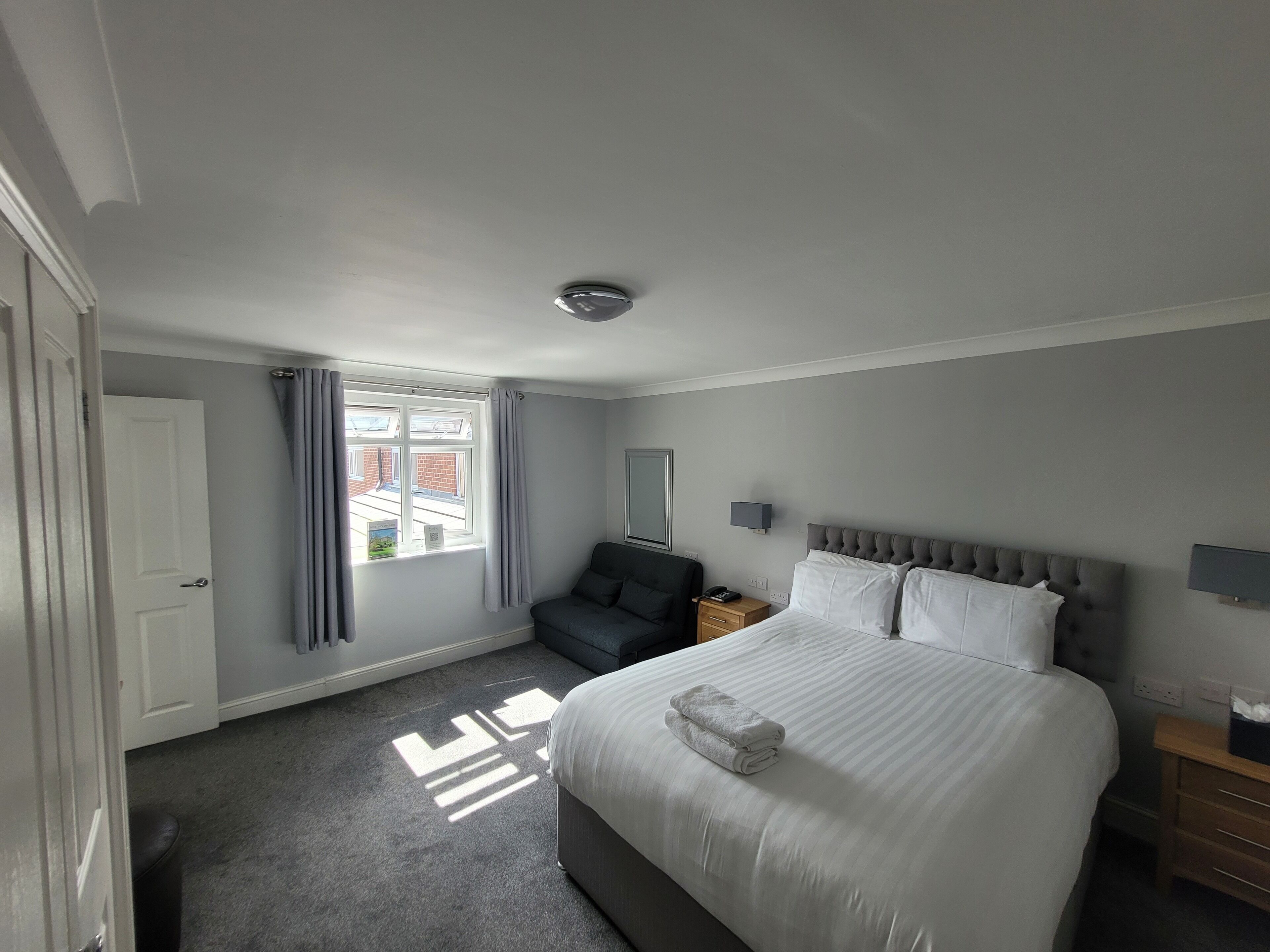 A hotel room with a large bed, a bedside table, a nightstand, a grey sofa, a window with curtains, and a view of buildings outside.