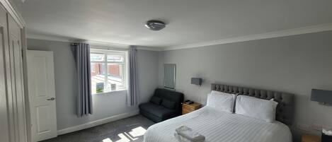 A hotel room with a large bed, a bedside table, a nightstand, a grey sofa, a window with curtains, and a view of buildings outside.