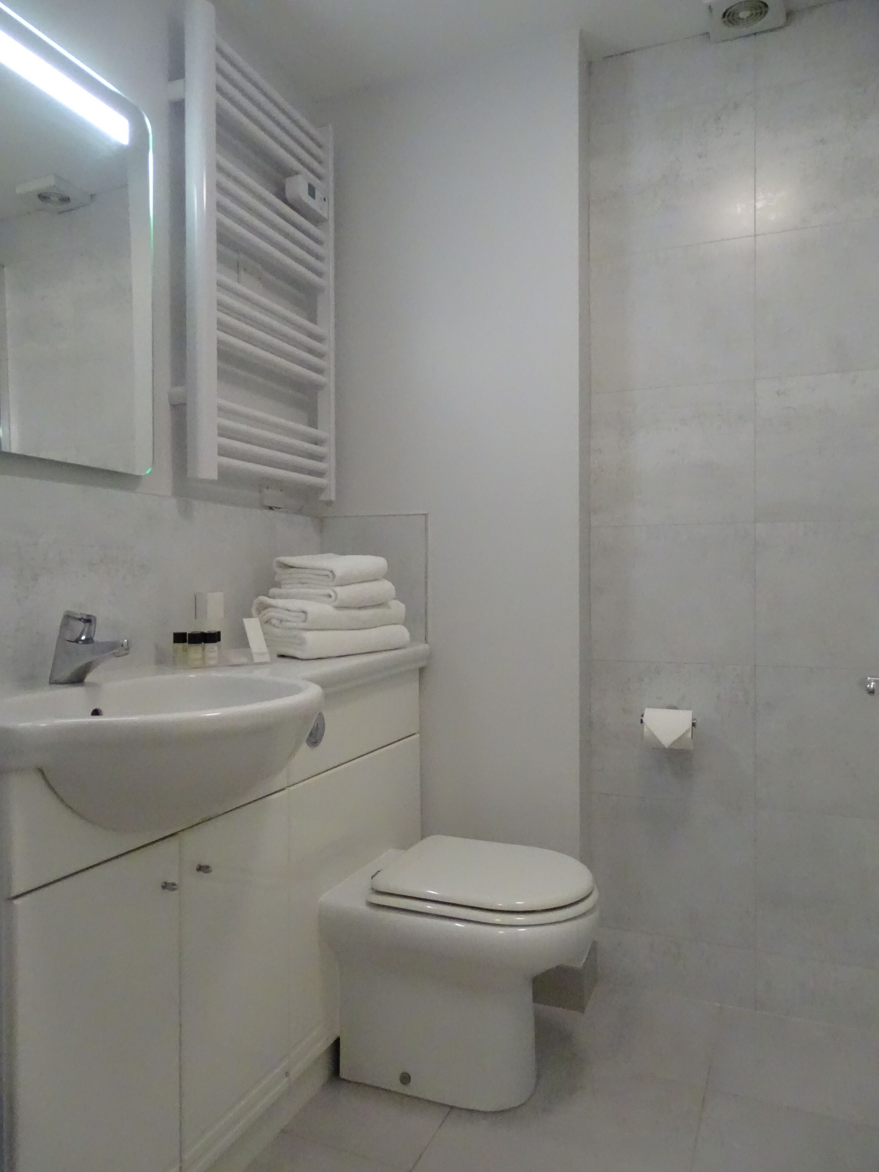 Et badeværelse med en hvid vask, spejl og toilet. Der er en håndklædestang over vasken og en toiletpapirholder på væggen.