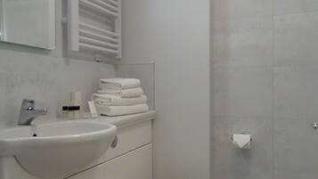 Un baño con lavamanos blanco, espejo y retrete. Hay un perchero sobre el lavamanos y un dispensador de papel higiénico en la pared.