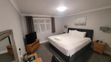 A hotel room with a large bed, a TV on a wooden stand, a desk with a chair, a nightstand, and a window with curtains.