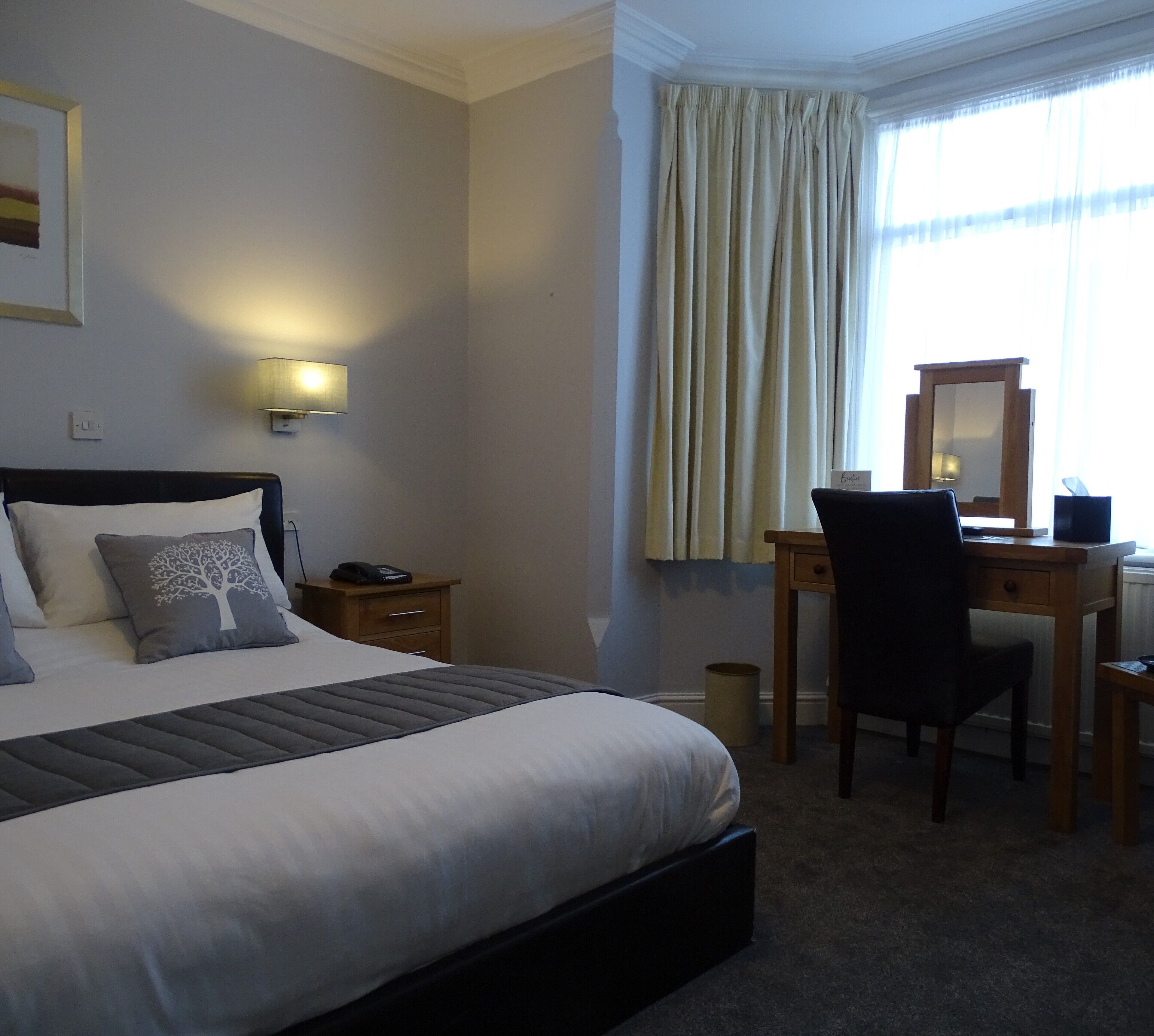 A hotel room with a bed, bedside table, lamp, desk, chair, and a window with curtains.