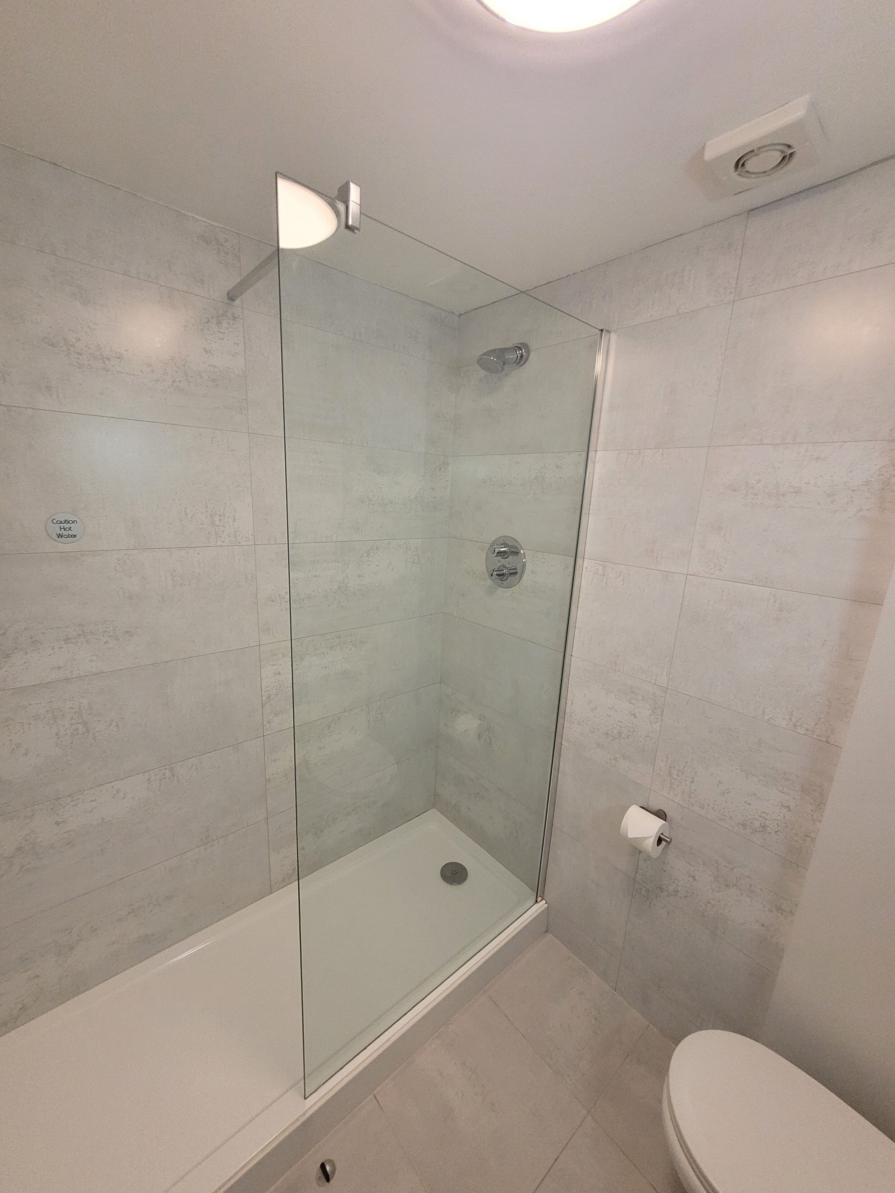 Une salle de bain moderne avec une douche à parois de verre, des toilettes et un panneau d’avertissement bien visible concernant l’eau chaude.