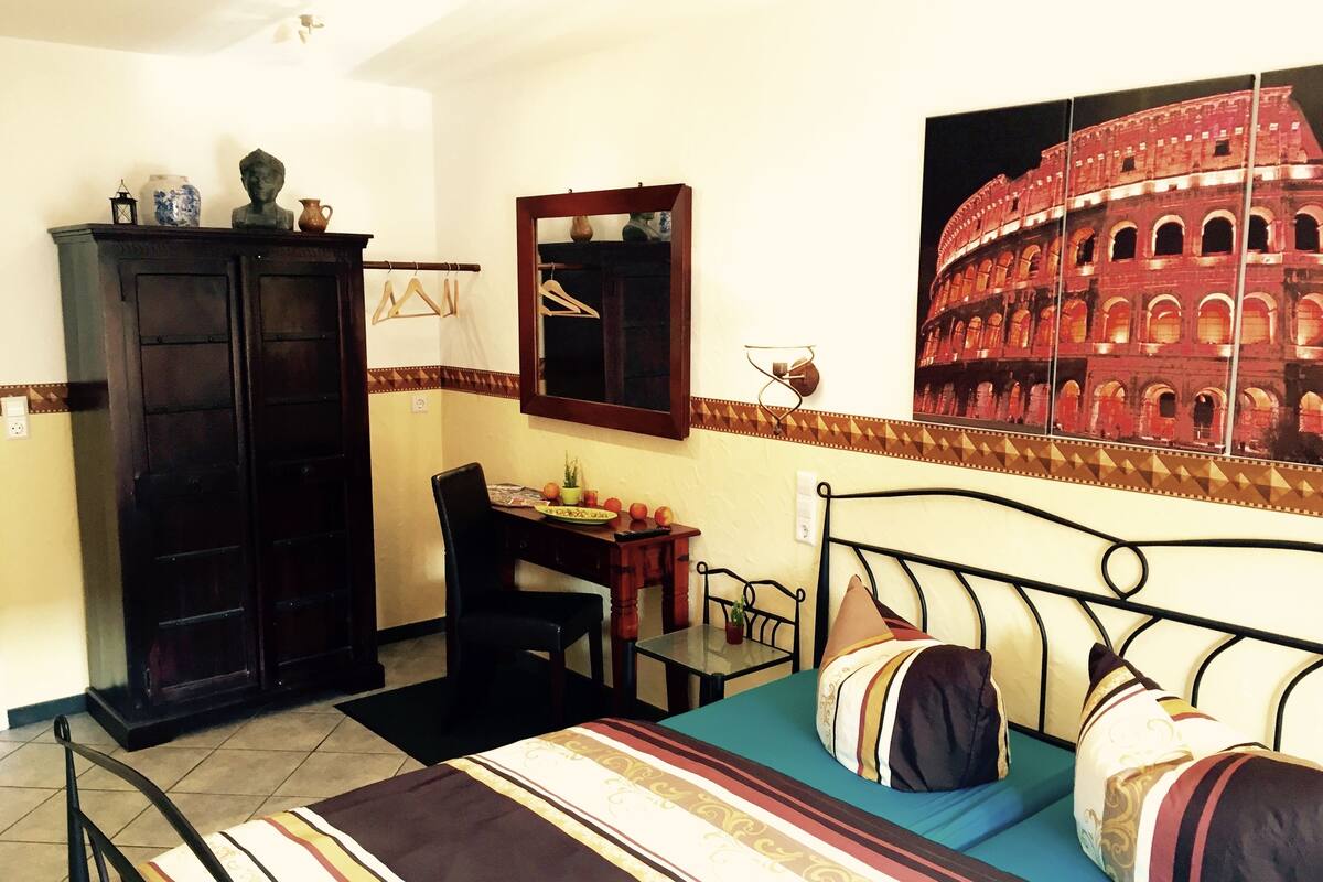 A room with a bed, a desk, a chair, a wardrobe, and a large wall painting of the Colosseum.