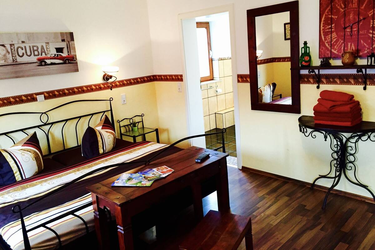 A room with a bed, a table, and a chair. There is a mirror on the wall and a shelf with items on it.