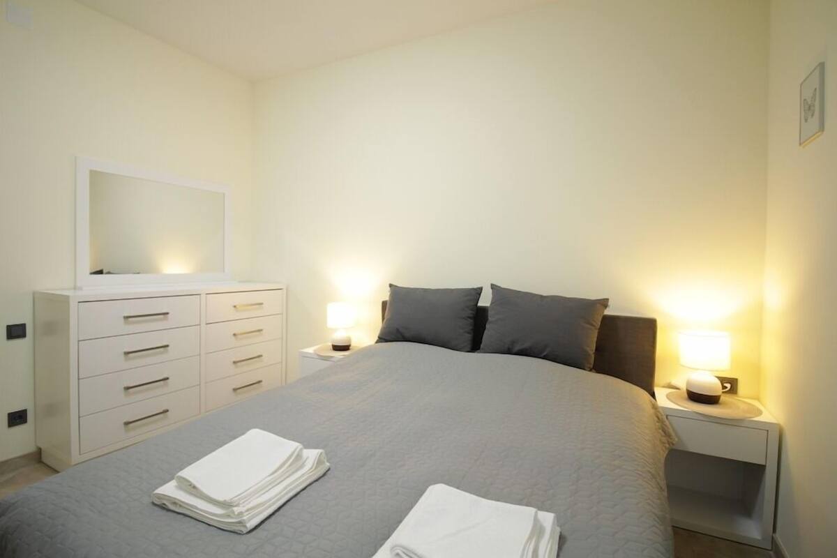 Un lit bien fait, recouvert d’une couvre-lit grise, avec deux oreillers et deux lampes de chevet. À gauche du lit, on trouve une commode blanche dotée de nombreux tiroirs.